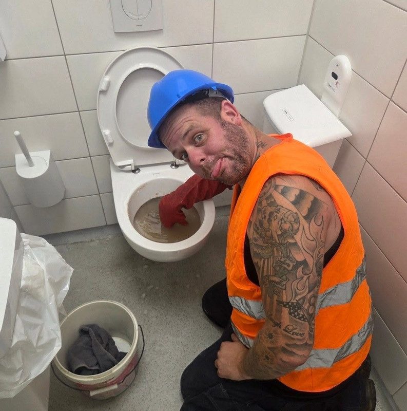 Danny met blauwe helm en oranje hesje, met zijn hand in een verstopte wc, trekt een gek gezicht